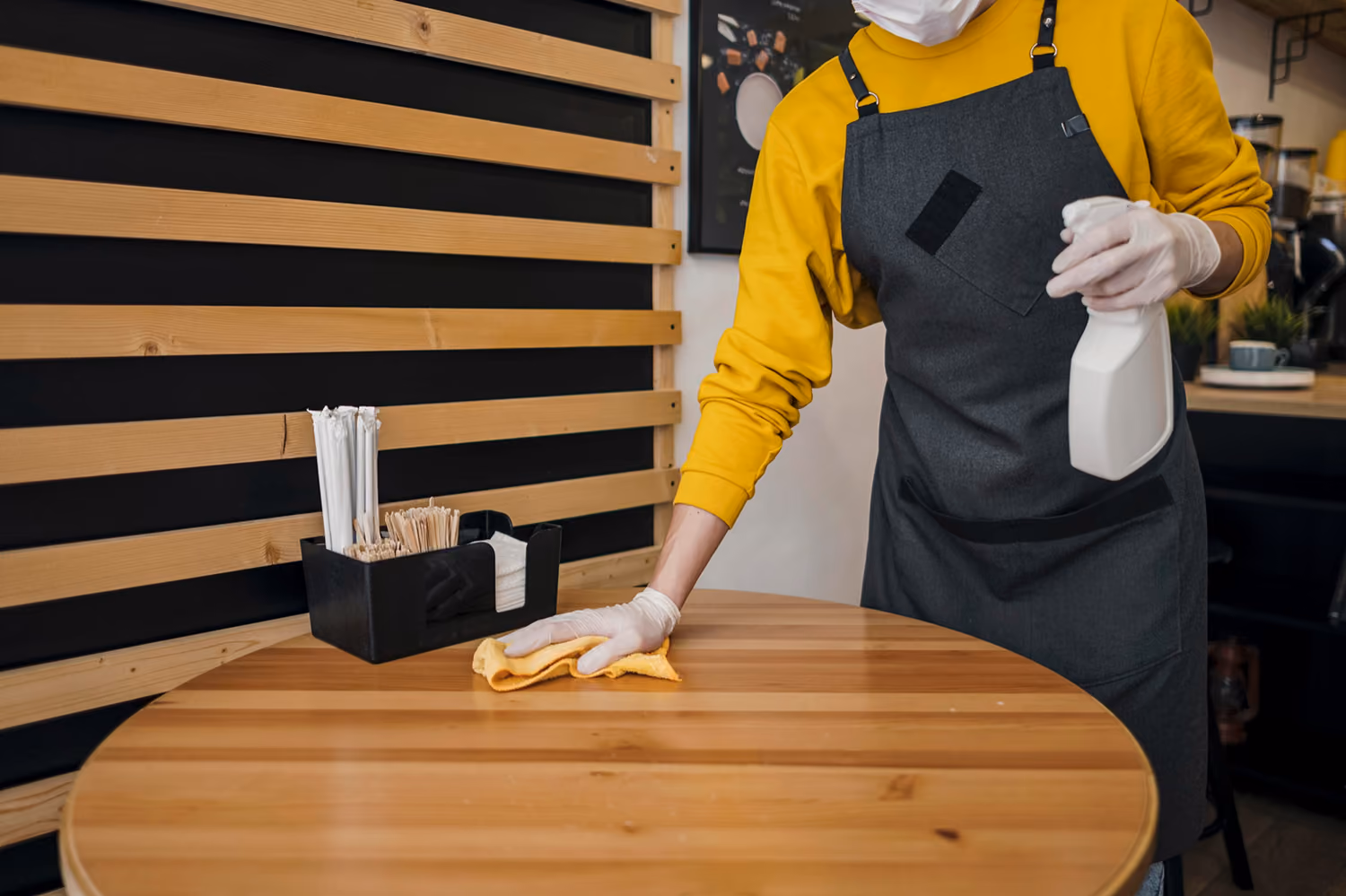 Gastronomiereinigung: Reinigungskraft in Firmenkleidung reinigt einen Holztisch in einem Café mit einem speziellen Reinigungsmittel und einem Tuch.