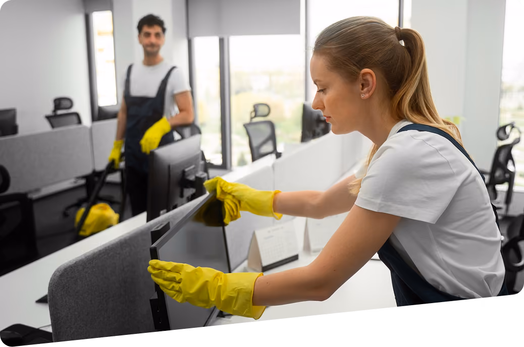 Unterhaltsreinigung für Unternehmen: Zwei Reinigungskräfte in Firmenkleidung reinigen ein Büro: Eine Mitarbeiterin wischt Staub vom Monitor, der Mitarbeiter saugt den Boden.