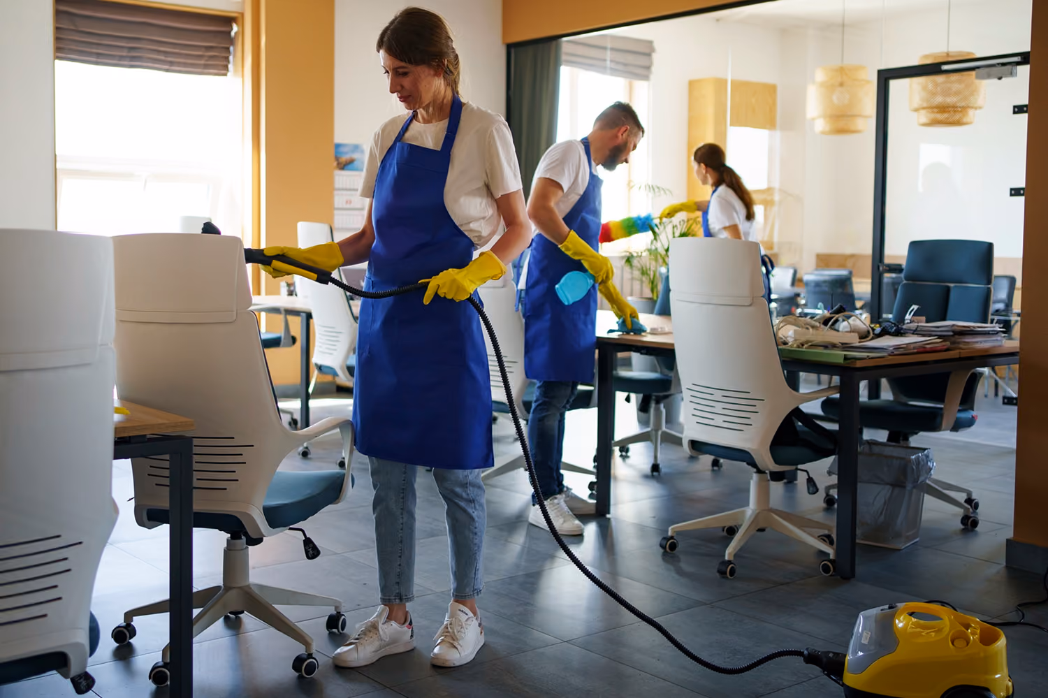 Unterhaltsreinigung für Unternehmen: Drei Reinigungskräfte in Firmenkleidung reinigen ein Büro: Der Mitarbeiter wischt Staub von einem Schreibtisch, eine Mitarbeiterin reinigt einen Sessel mit einem speziellen Staubsauger, die dritte Mitarbeiterin gießt die Blumen.