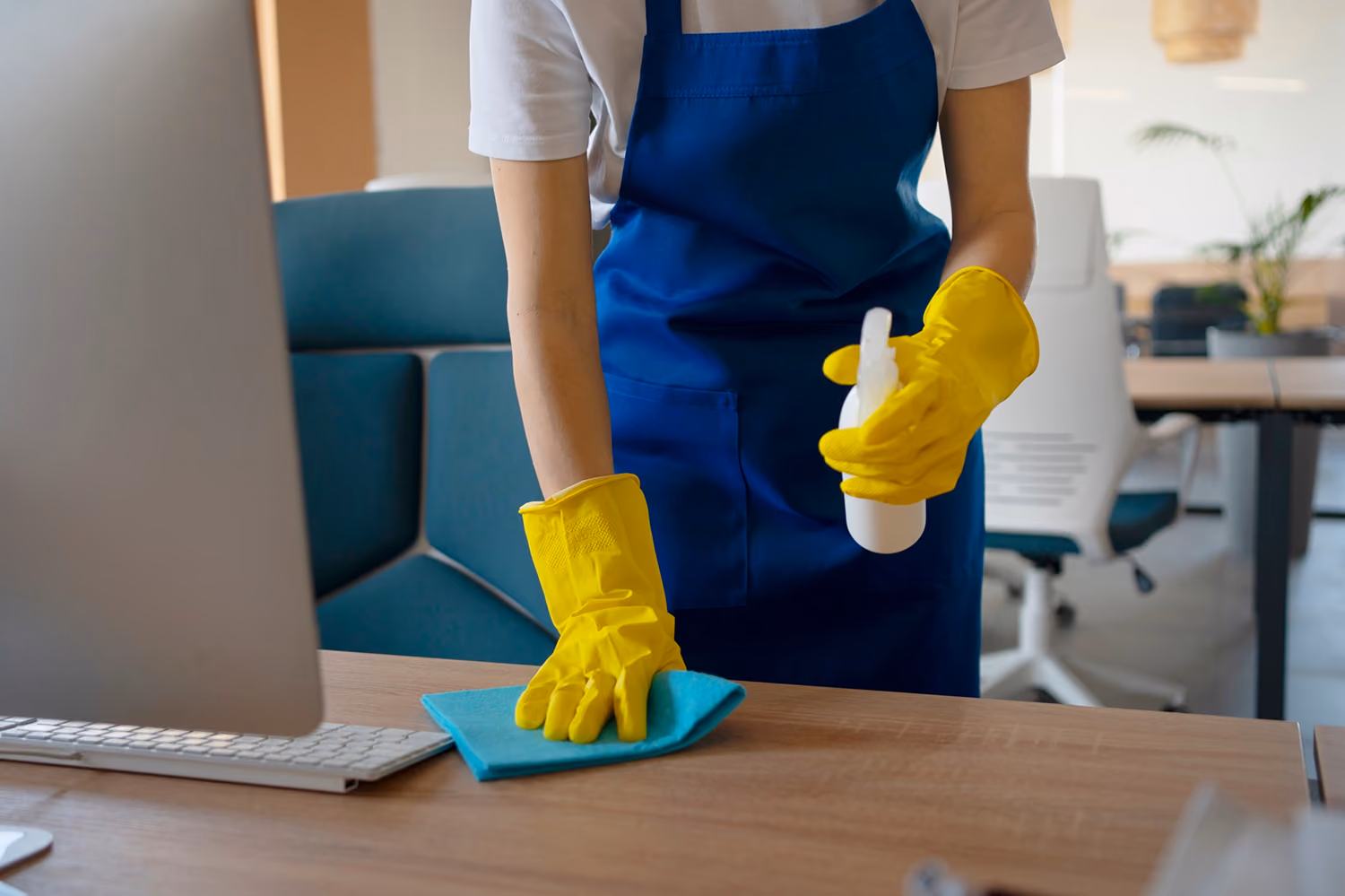 Büroreinigung: Nahaufnahme einer Reinigungskraft in Firmenkleidung, die eine Schreibtischoberfläche im Büro mit einem speziellen Reinigungsmittel und einem Tuch reinigt.