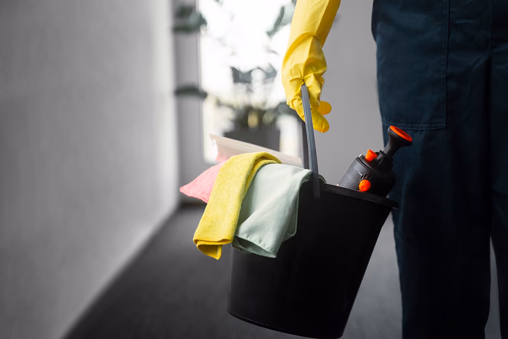 Individuelle Reinigungsleistungen: Eine Reinigungskraft in dunkelblauer Arbeitskleidung trägt einen schwarzen Reinigungseimer mit gelben Handschuhen, Reinigungstüchern und Reinigungsmitteln durch einen Flur.