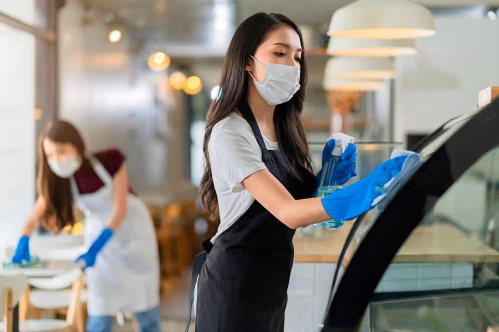 Gastronomiereinigung: Zwei Reinigungskräfte in Arbeitskleidung reinigen einen gastronomischen Bereich: Eine Mitarbeiterin reinigt eine gebogene Glasvitrine mit ausgestellten Waren, die zweite wischt einen Tisch für Gäste.