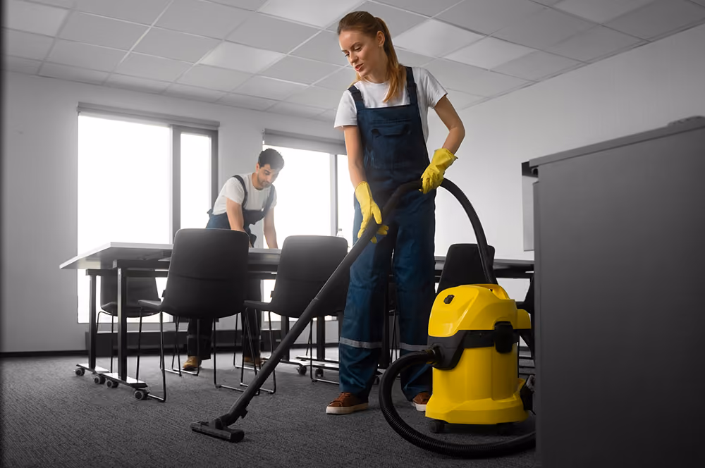 Büroreinigung: Zwei Reinigungskräfte in Firmenkleidung reinigen ein Büro: Die Mitarbeiterin reinigt den Boden mit einem Waschsauger, der Mitarbeiter wischt Staub von einem Konferenztisch.