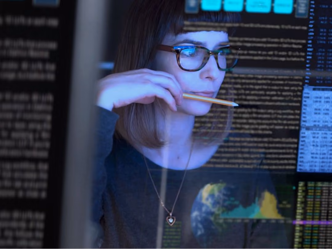 Young woman with glasses holding a pencil, focused on data and code on transparent screens.
