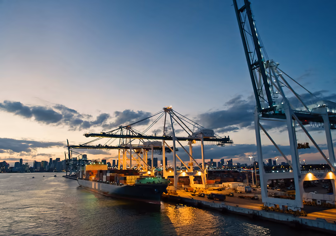Cargo ship docked at port with cranes unloading containers at sunset with city skyline in the background.
