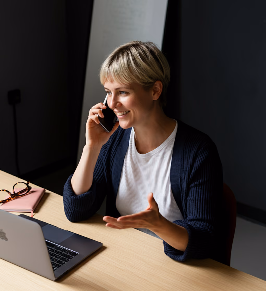 Smiling woman in a dark cardigan talking on phone at desk with laptop and glasses.