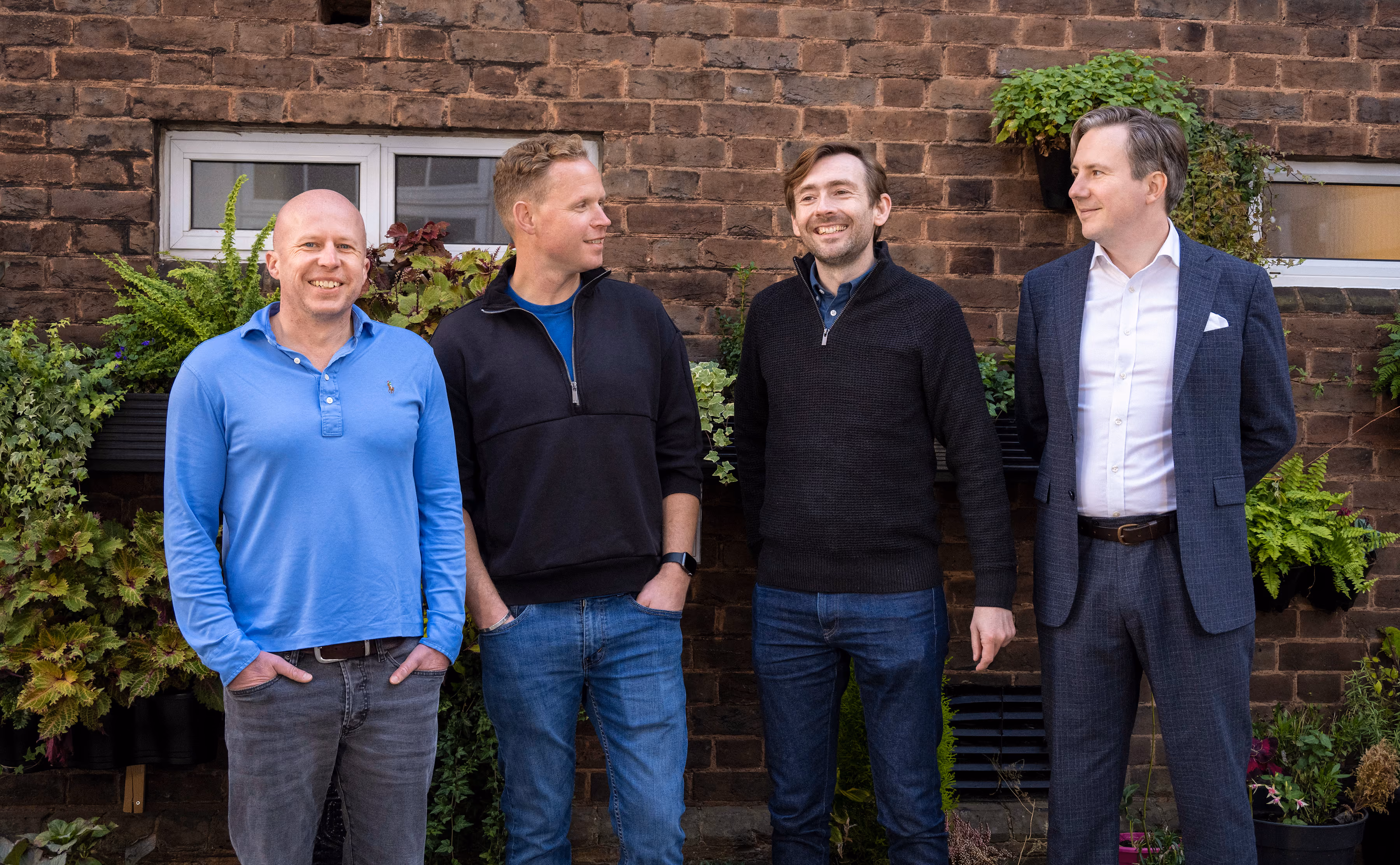 TreasurySpring's four founders (Henry Adams, Kevin Cook, Matthew Longhurst, and James Skillen) standing side by side smiling outdoors against a brick wall with plants and windows.