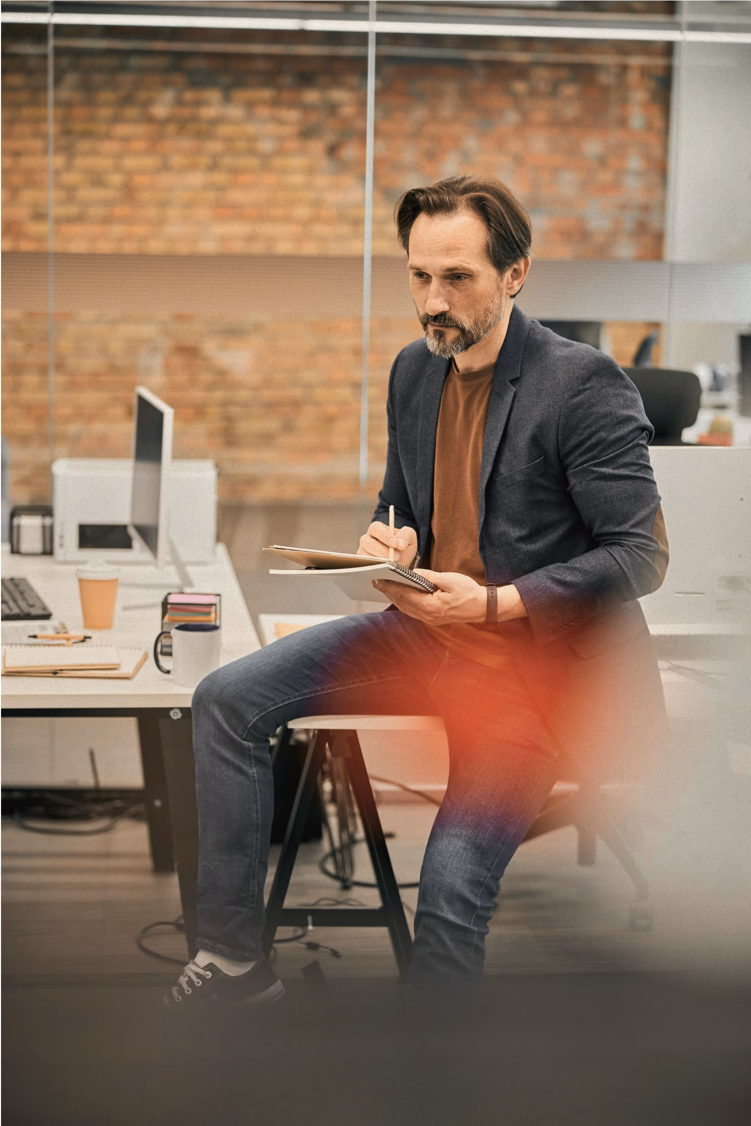 Man in blazer and jeans sitting on a desk, writing in a notebook in an office with brick wall.