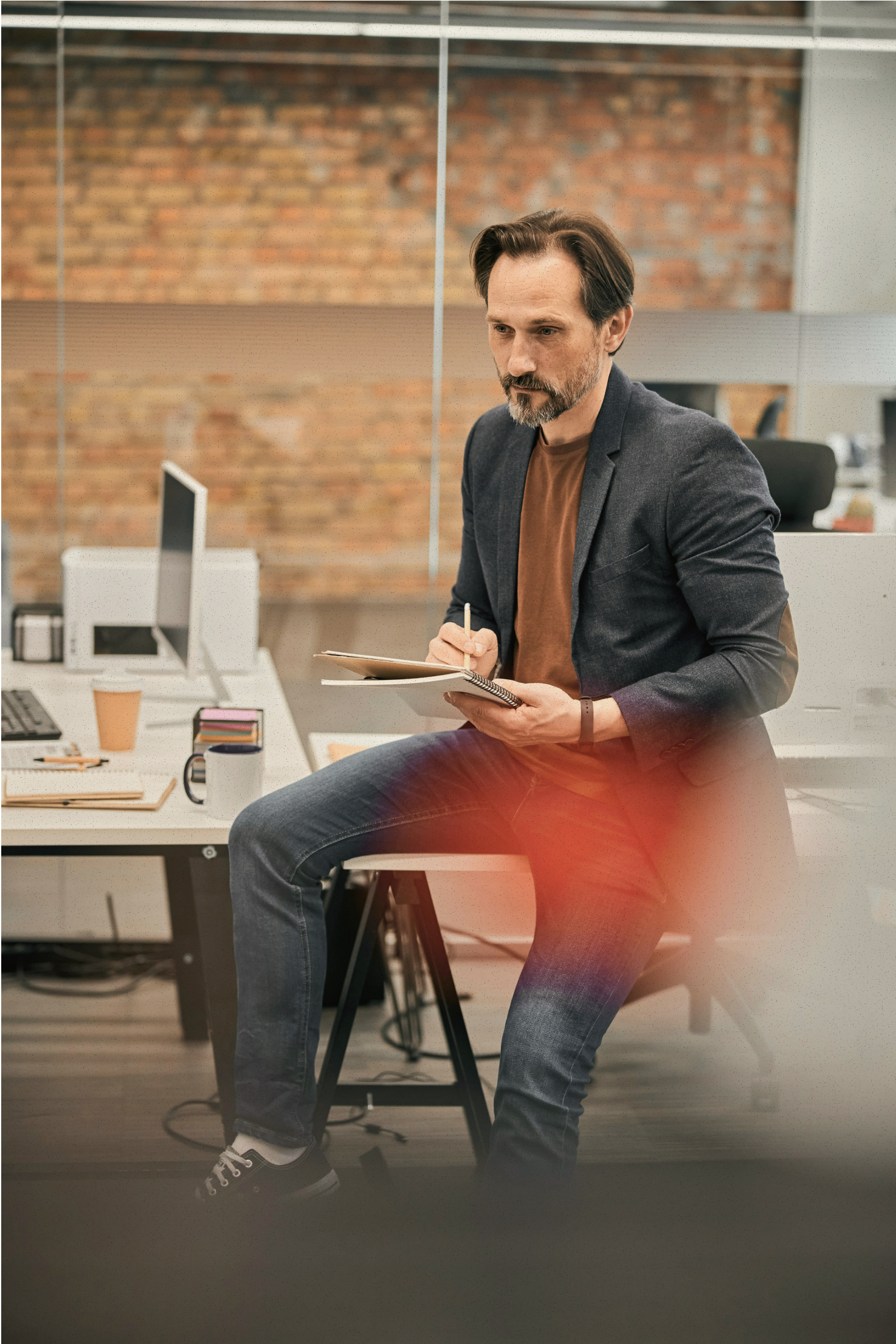 Man in blazer and jeans sitting on a desk, writing in a notebook in an office with brick wall.