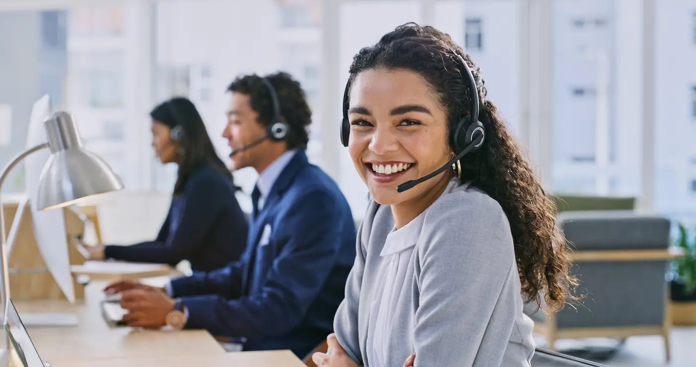 Smiling woman wearing headset in office with two coworkers using headsets at computers in background.
