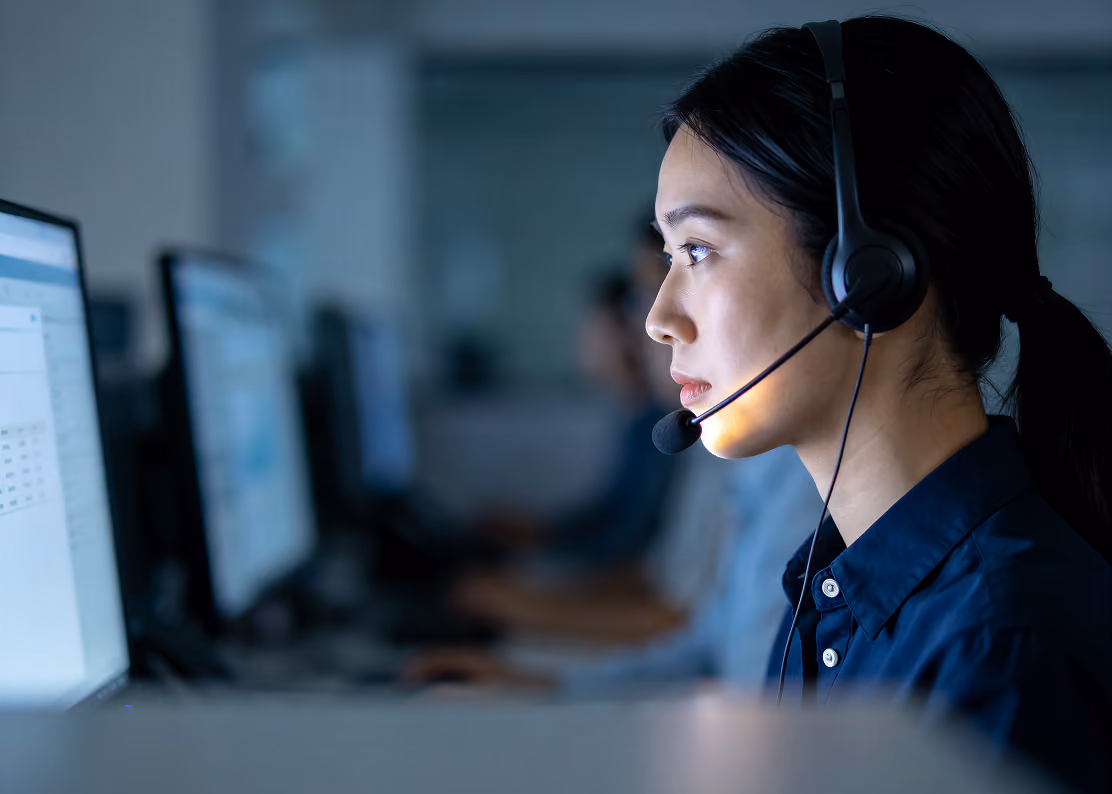 Woman wearing headset working at computer in a dimly lit office with other workers in background.