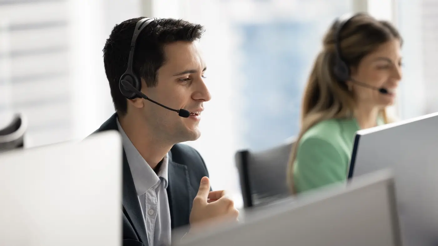 Two call center agents wearing headsets working at computers in a bright office.