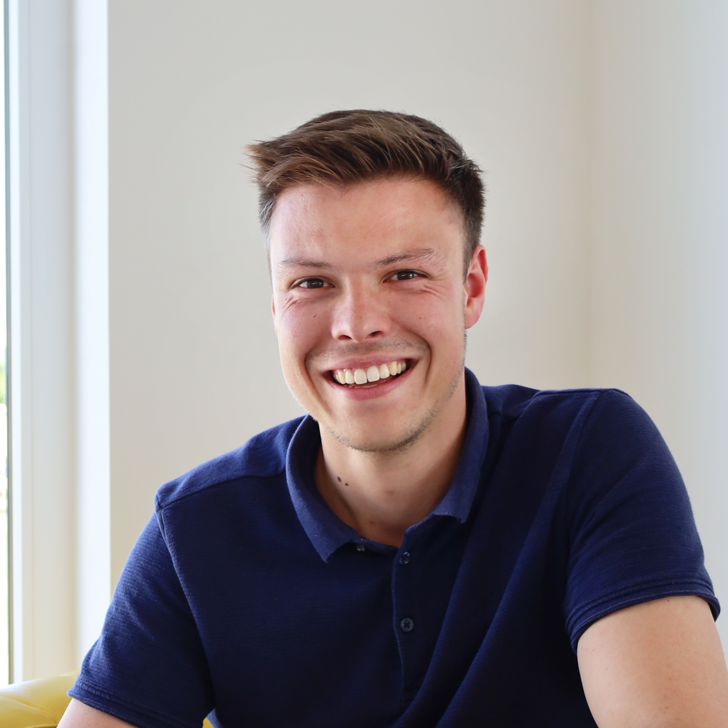 Junger Mann in dunkelblauem Poloshirt lächelt in hellen Innenraum neben Fenster.