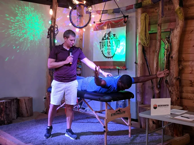 Therapist assists man lying face down on massage table with arms extended in a rustic room.