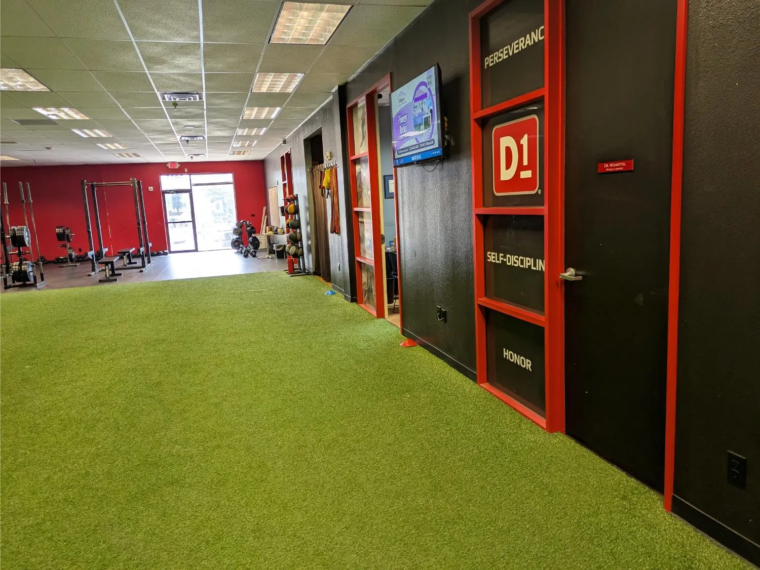 View of a gym interior with green turf, weight racks, and a black and red wall with motivational words.