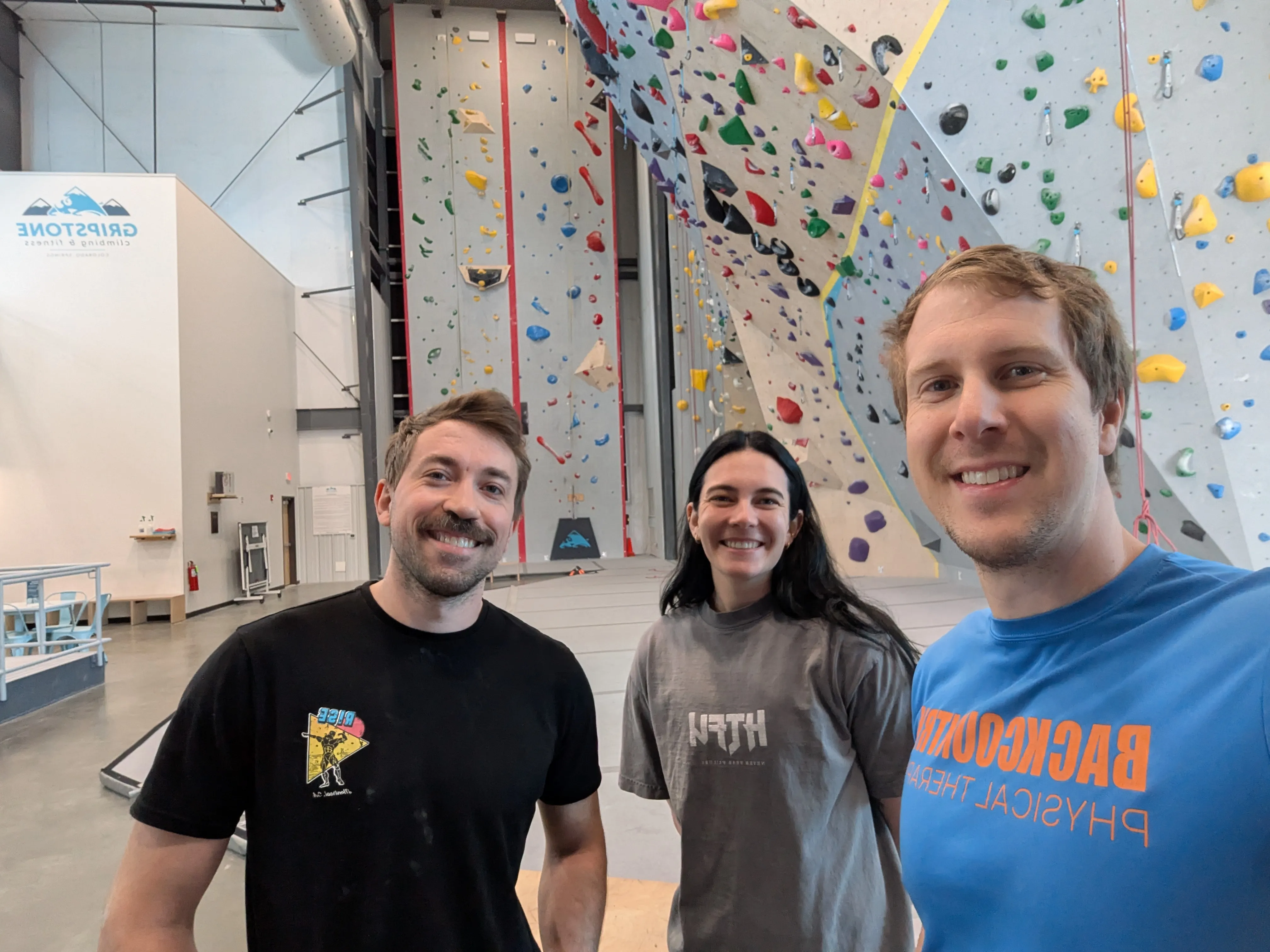 Three smiling people posing inside an indoor rock climbing gym with colorful climbing holds on the walls.