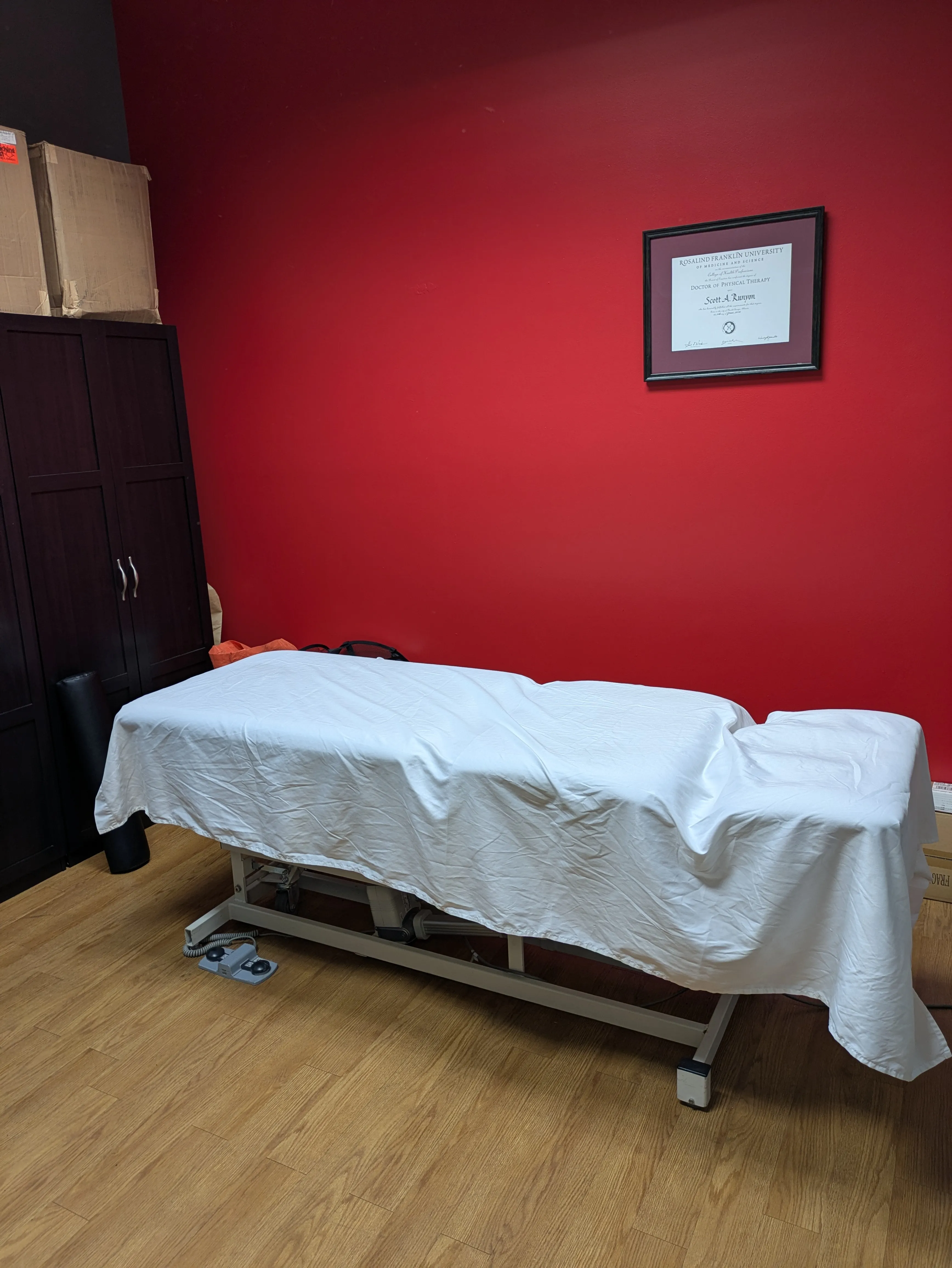 Massage or therapy table covered with a white sheet in a room with red wall and wooden floor.