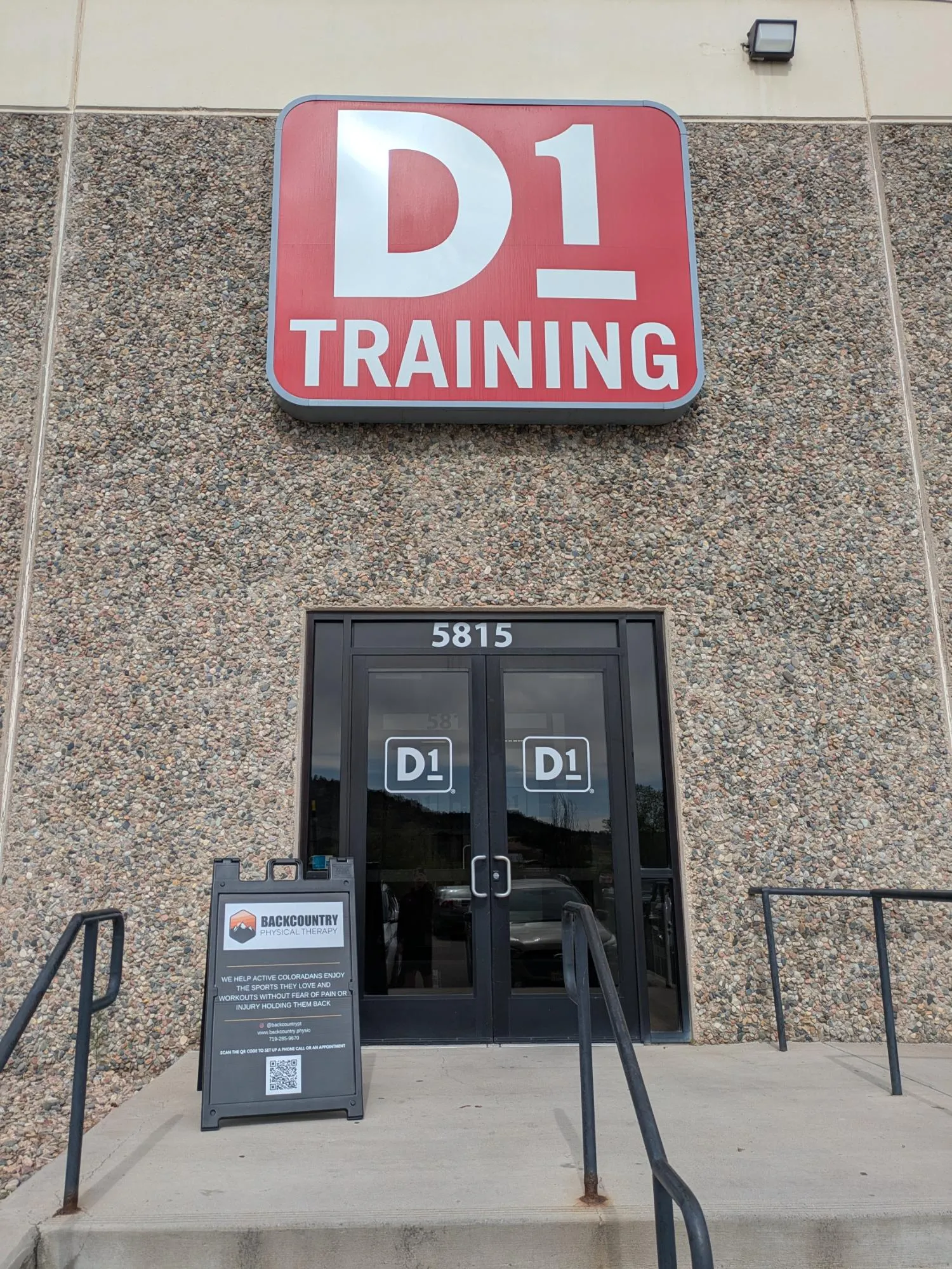 Entrance of D1 Training gym with signage and Backcountry Physical Therapy sign on sidewalk.