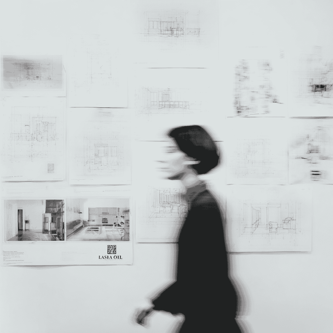 Blurred person walking past a white wall with architectural sketches and photos pinned on it.