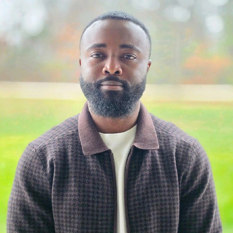 Portrait of a man with beard wearing a brown jacket and white shirt standing outdoors.