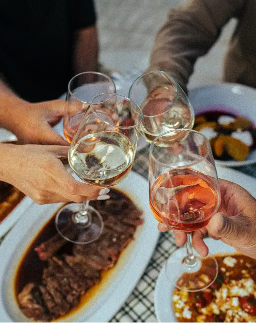 Four people clinking glasses of white and rosé wine over a table with dishes including steak and pizza.