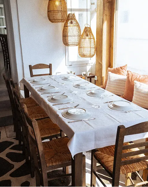Dining table set for six with white cloth and glasses under hanging wicker lamps in bright room.