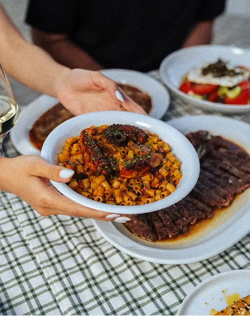 Hands holding a plate of pasta topped with grilled octopus over a table with various dishes.
