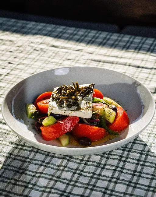 Greek salad with tomatoes, cucumbers, olives, capers, and feta cheese topped with herbs in a white bowl.