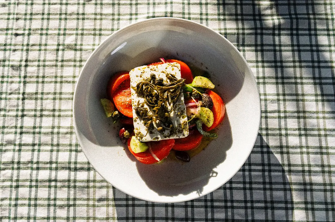 White bowl with sliced tomatoes, cucumbers, olives, feta cheese, and herbs on checked tablecloth.