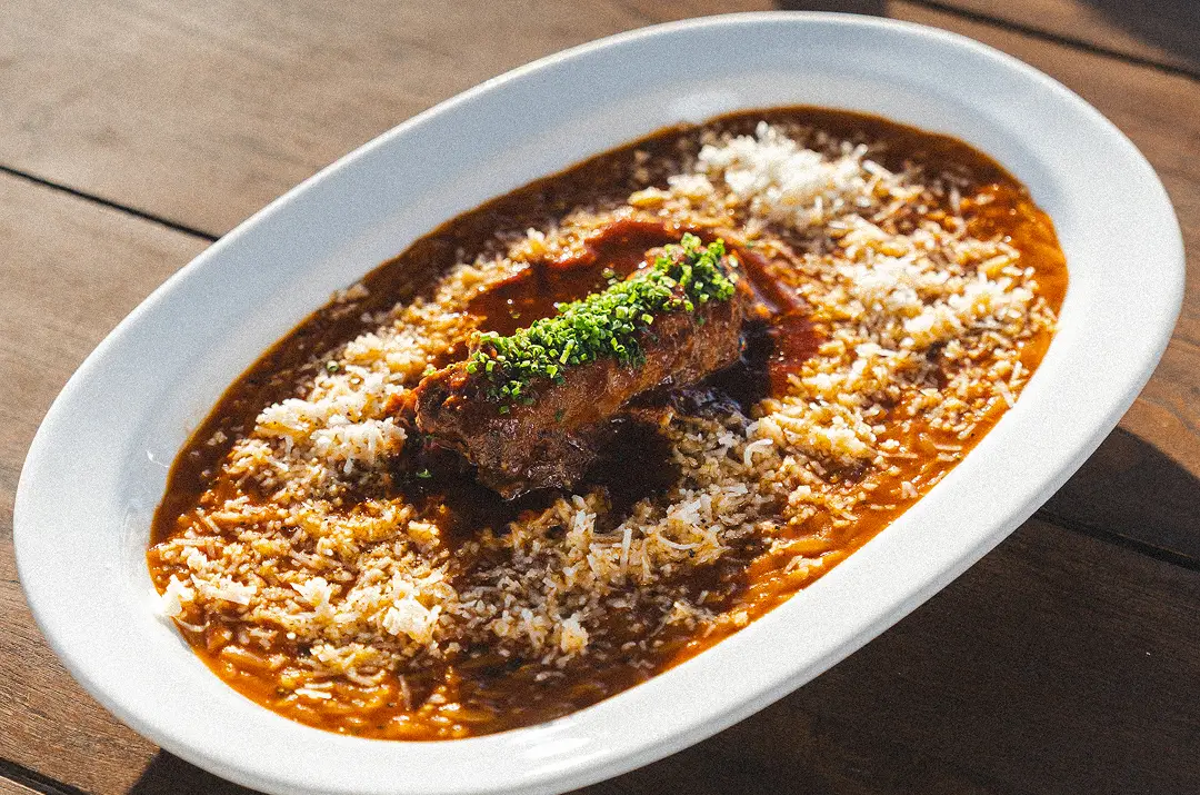 Plate of rice and sauce topped with a piece of meat garnished with chopped herbs on wooden table.