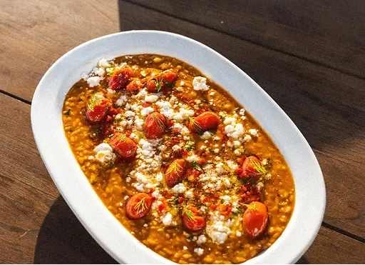 White dish of lentil stew garnished with halved cherry tomatoes, herbs, and crumbled cheese on wood table.