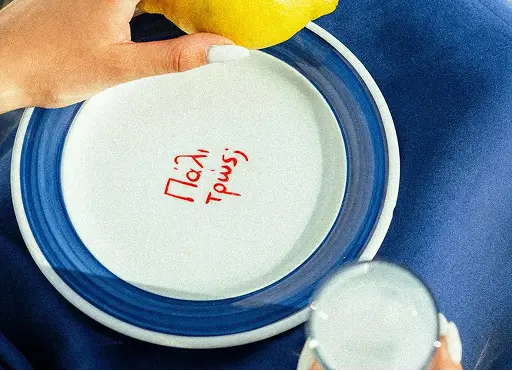 Hand squeezing lemon over plate with Greek text and glass of iced drink on blue surface.