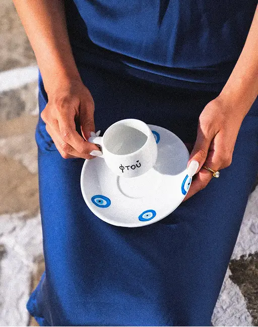 Person in blue dress holding white cup and saucer decorated with blue eye symbols.