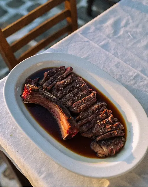 Sliced grilled steak with bone served on a white plate with dark sauce on a tablecloth.