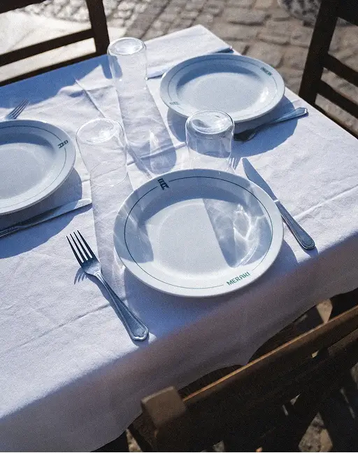 Outdoor dining table set with white plates, clear glasses, forks, knives, and white tablecloth in sunlight.