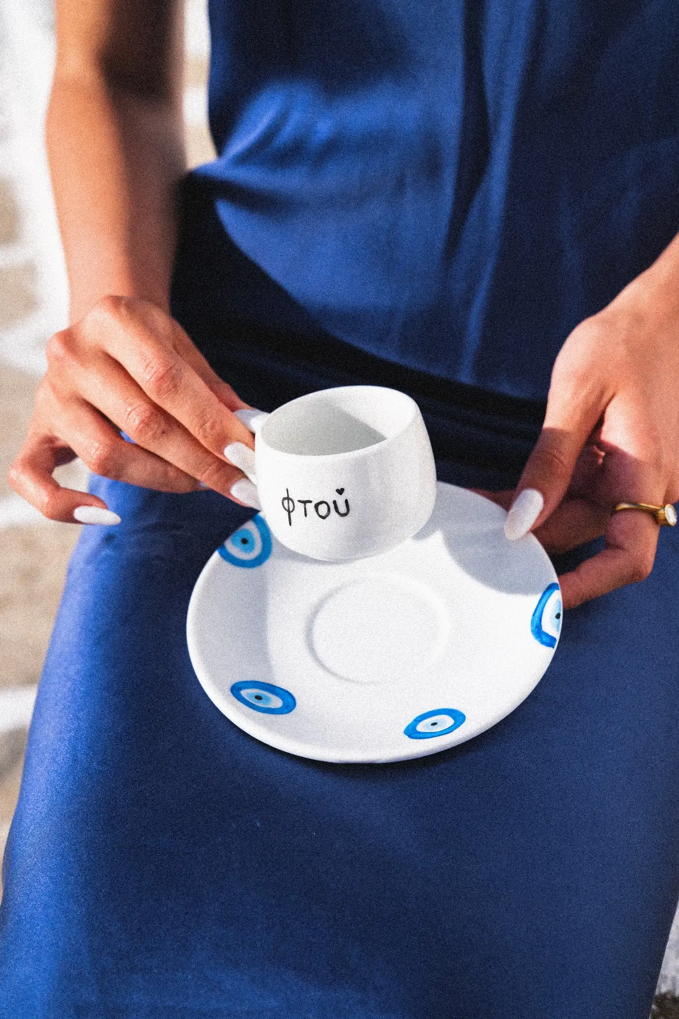 Person holding a white cup with Greek text and a saucer decorated with blue evil eye symbols.