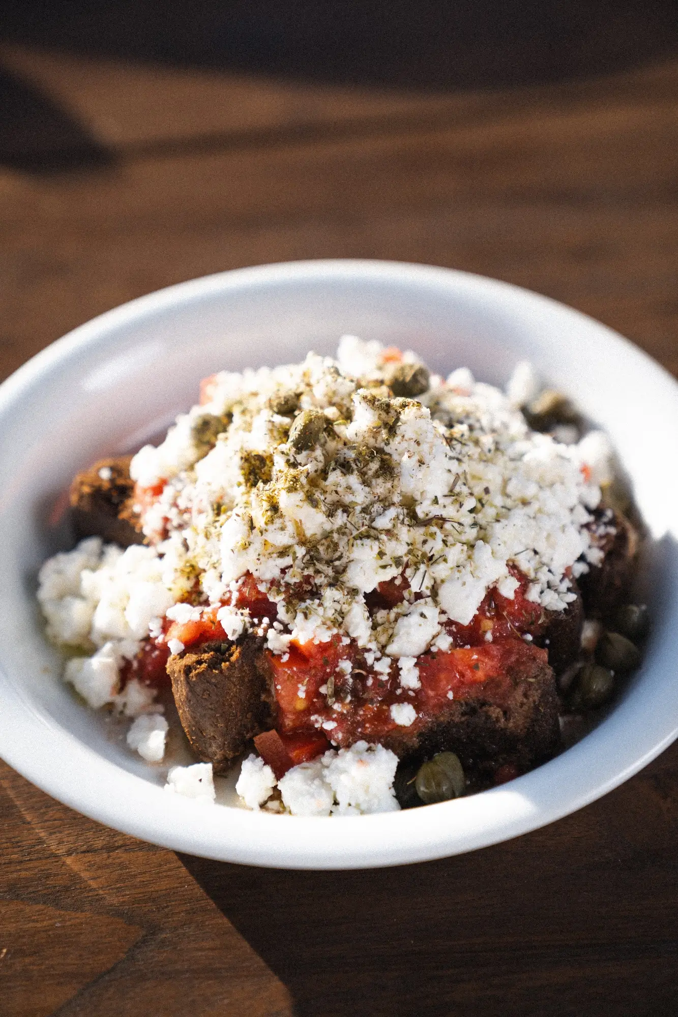 White bowl with Greek-style salad topped with feta cheese, tomato sauce, herbs, and capers.