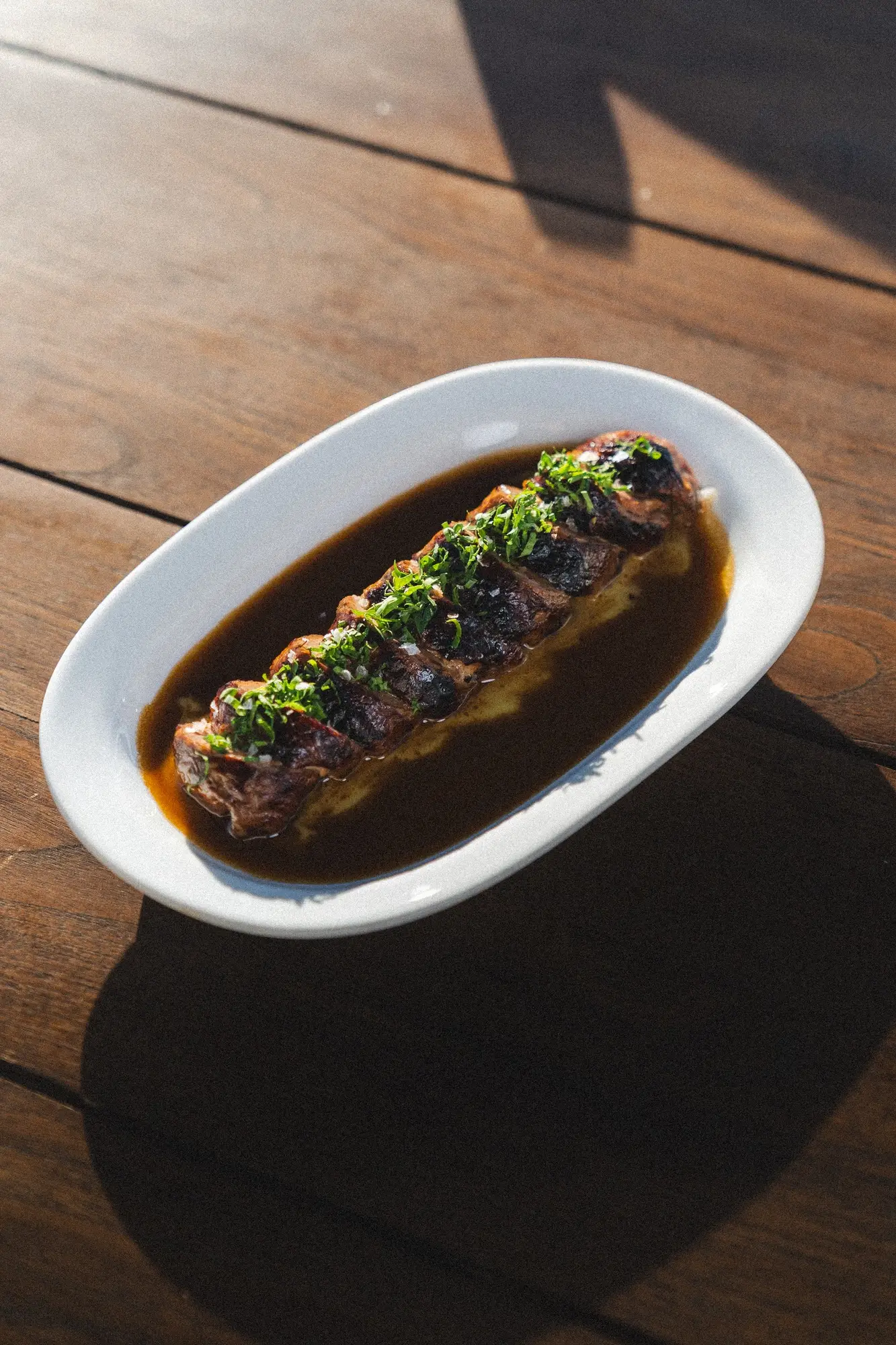 Grilled sliced steak topped with chopped herbs served in dark sauce on white oval plate.