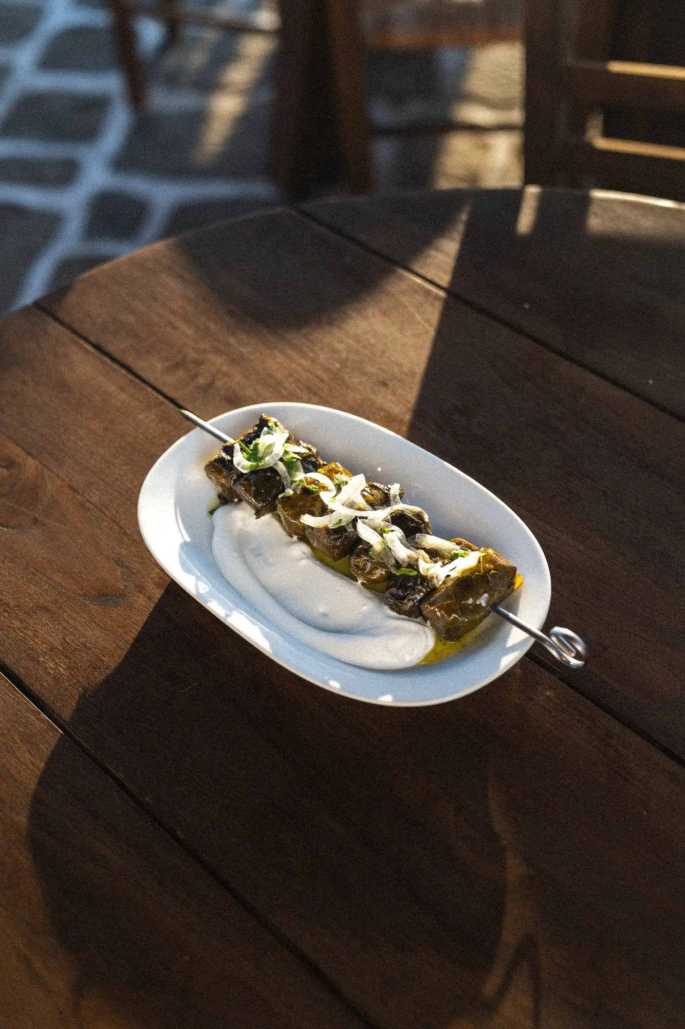 Grilled stuffed grape leaves on a skewer with creamy sauce and chopped onions on a white plate.