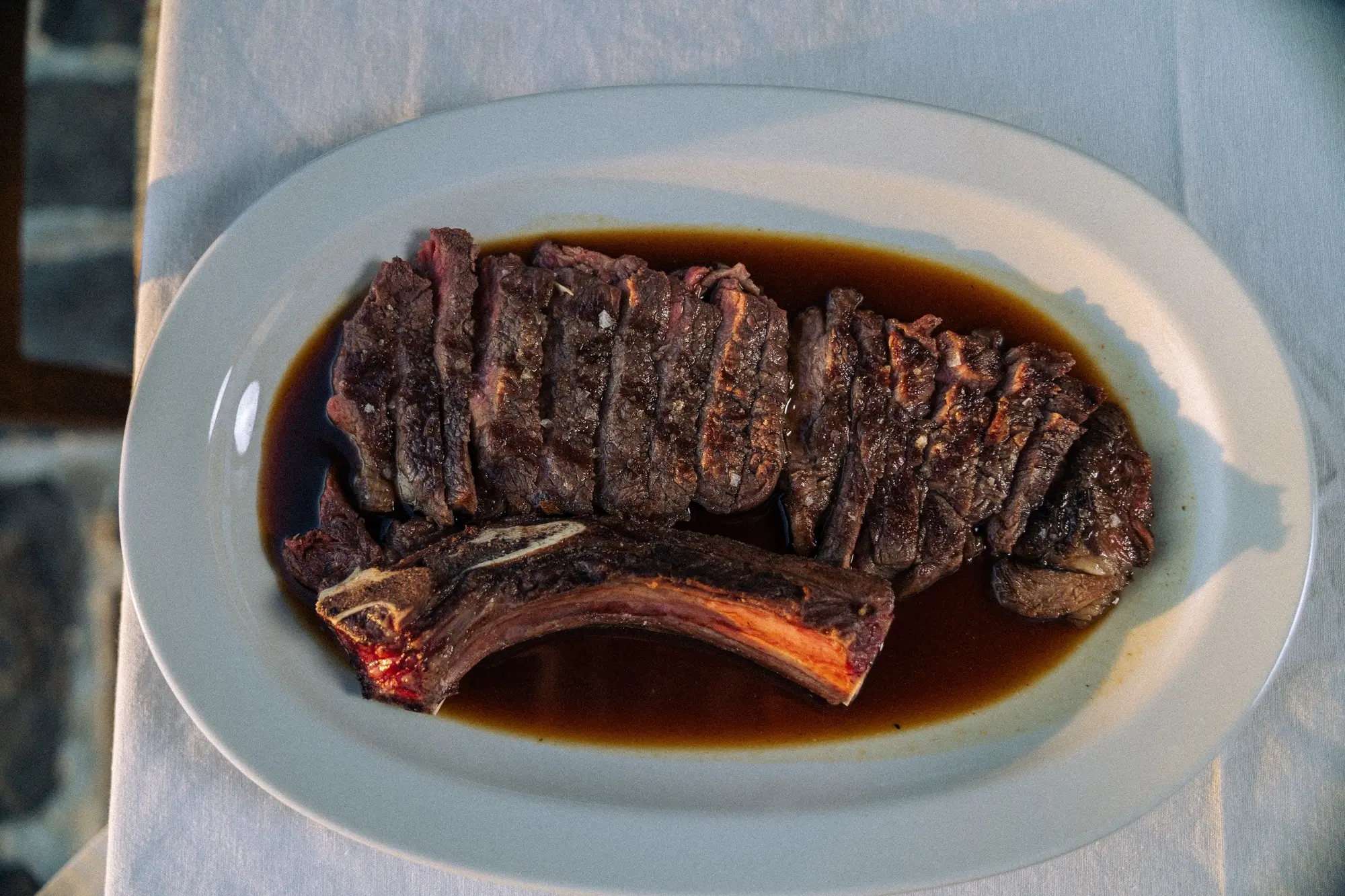 Grilled sliced steak with a bone on a white plate with dark sauce on a white tablecloth.
