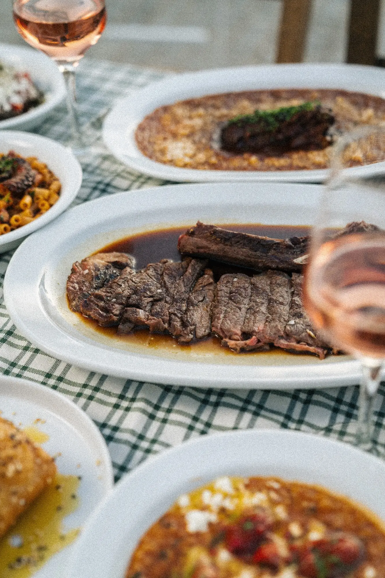 Plated grilled steak with sauce, surrounded by other dishes and glasses of rosé wine on a checkered tablecloth.