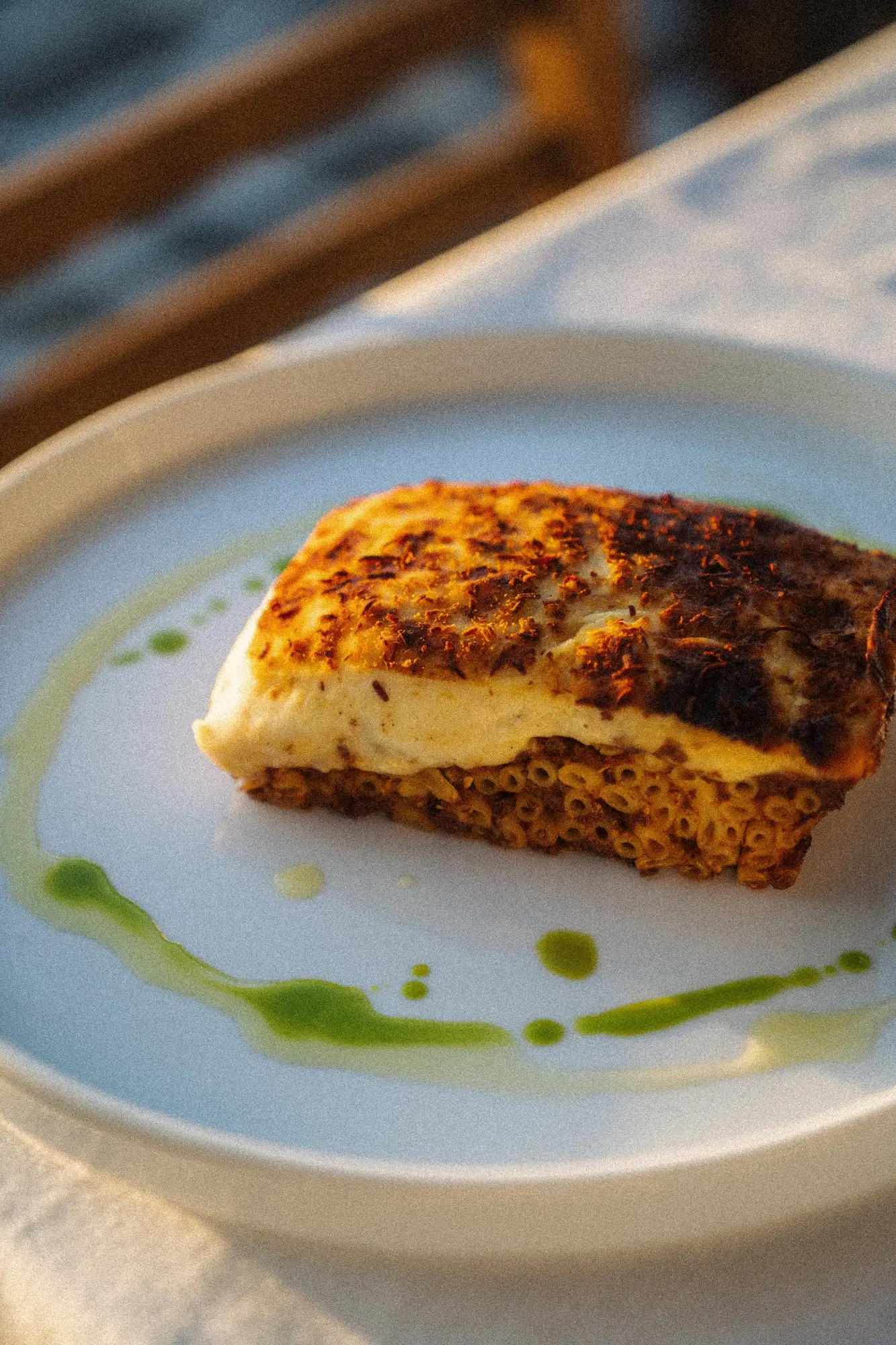 Baked pasta dish topped with golden grilled cheese and green oil drizzle on white plate.