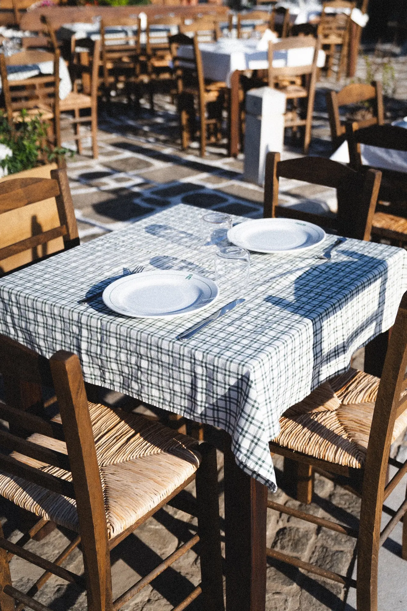 Outdoor restaurant table with checkered cloth, two plates, glasses, and wooden chairs on stone floor.