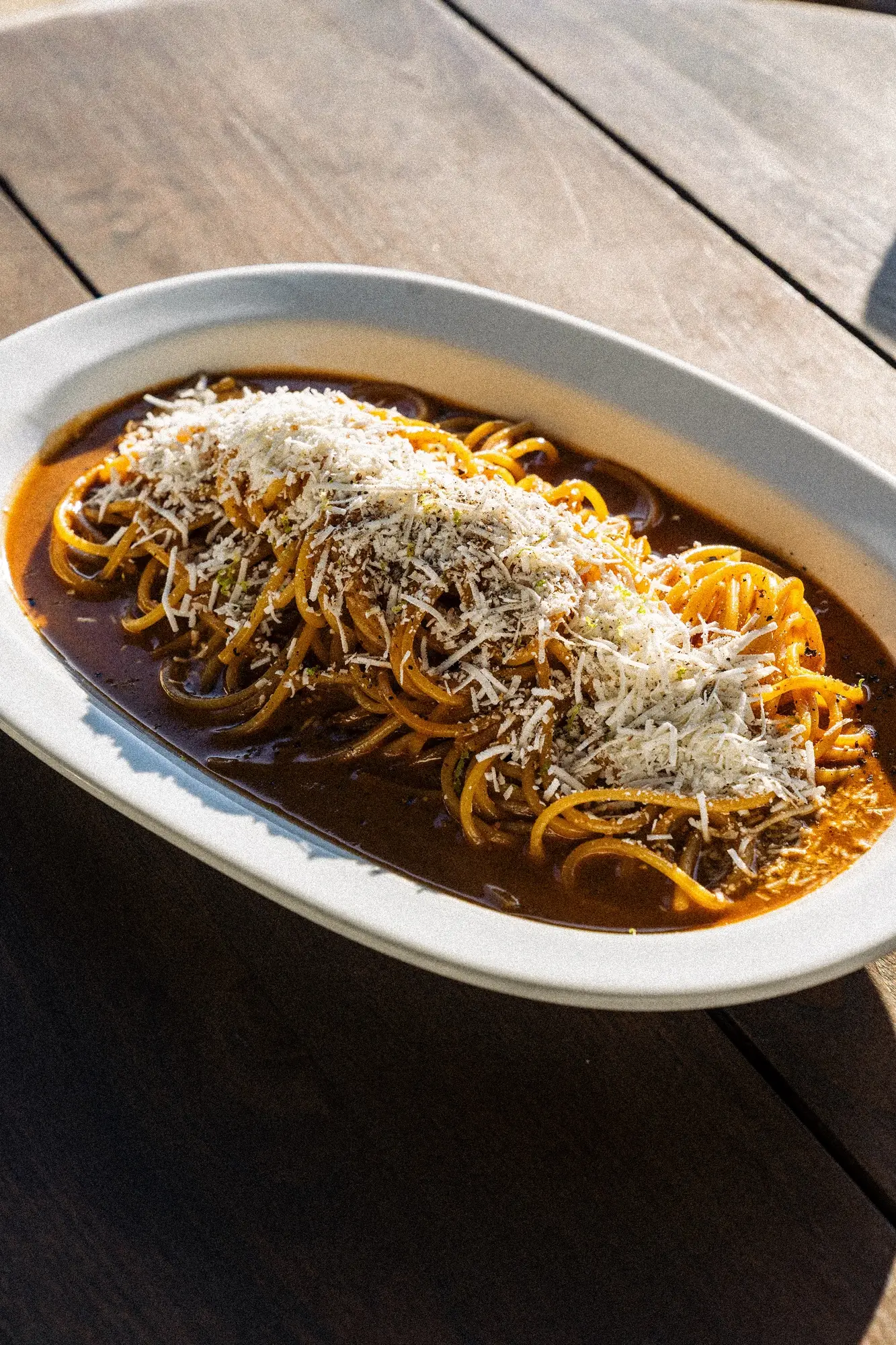 Plate of spaghetti with dark sauce topped with grated cheese on a wooden table.