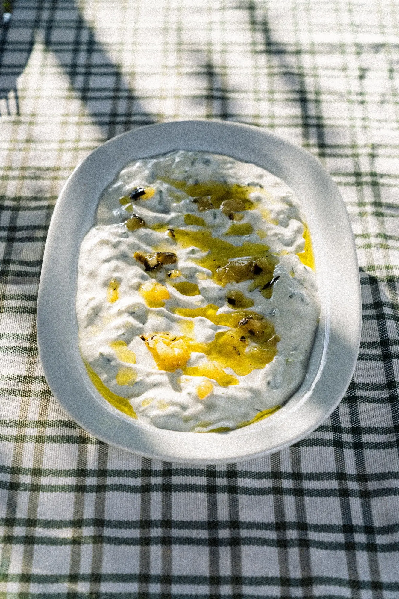 White dish with creamy yogurt dip topped with olive oil and herbs on a green checkered cloth.
