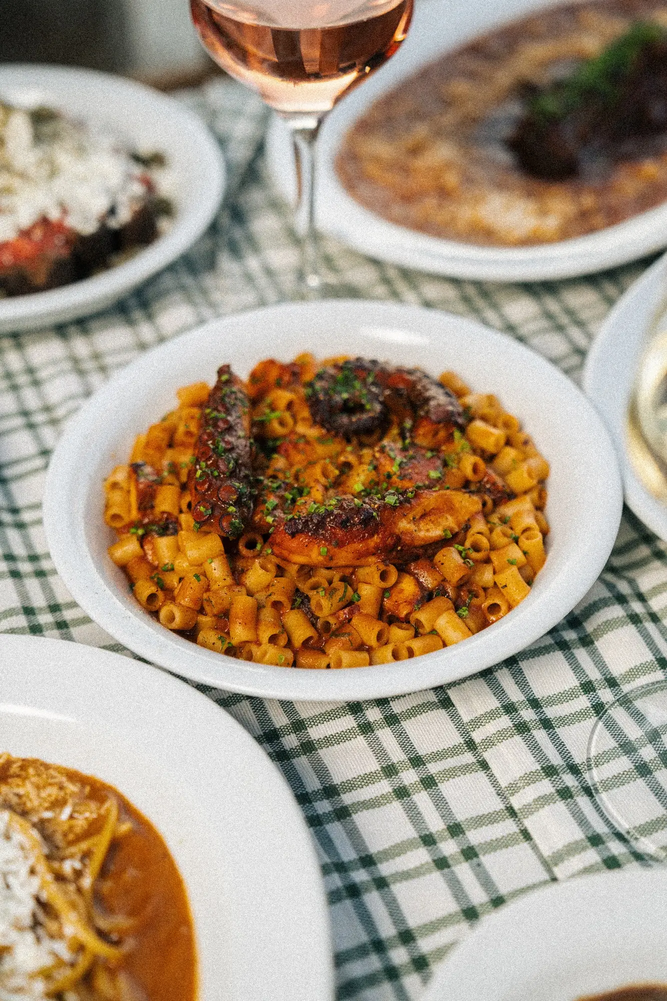 White bowl of pasta with grilled octopus tentacles garnished with herbs on a plaid tablecloth.