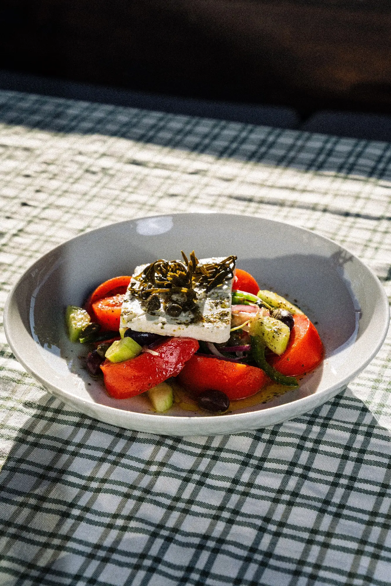 Greek salad with tomatoes, cucumbers, olives, feta cheese, and herbs on a checked tablecloth.