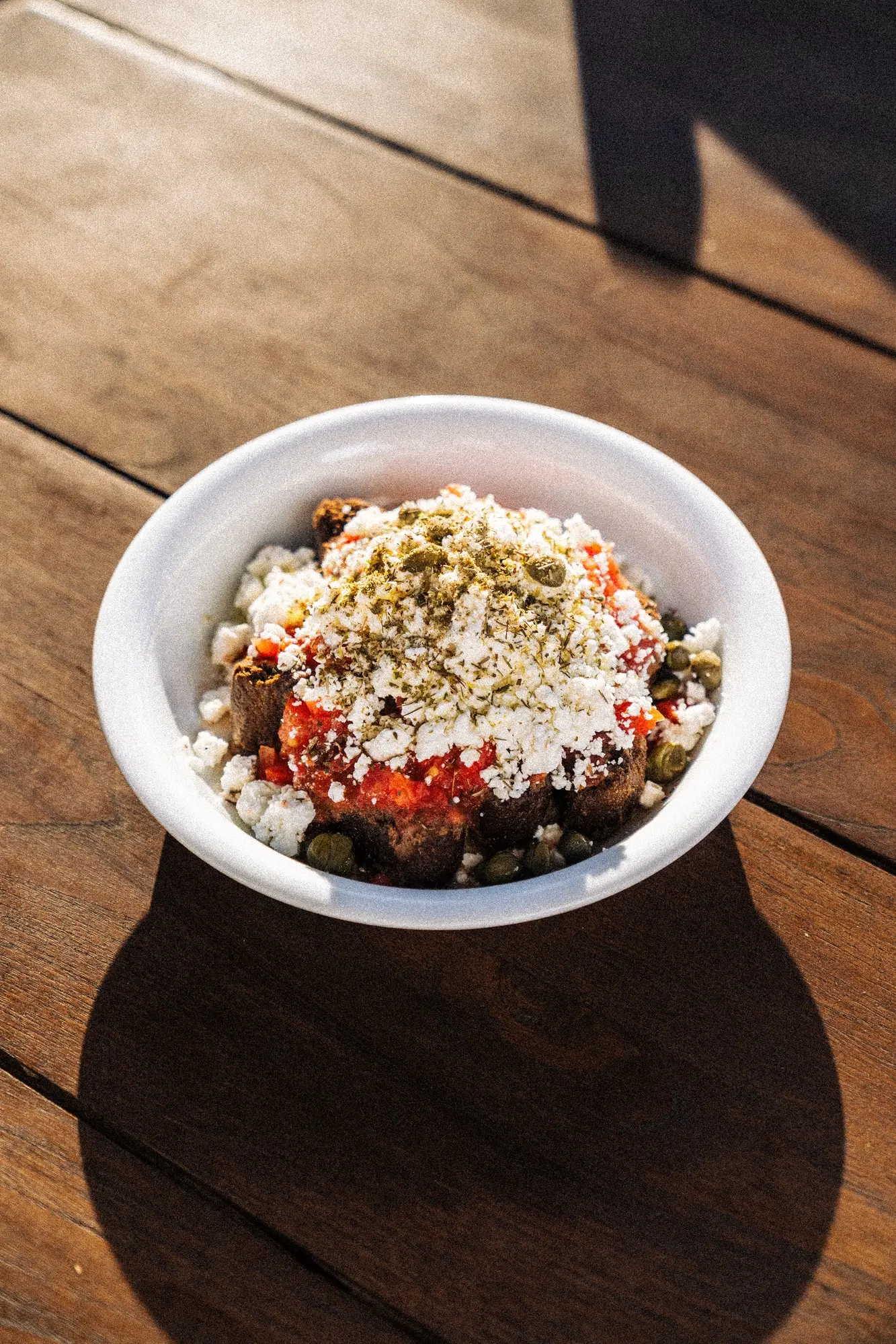 White bowl with bread, tomato sauce, cheese, herbs, and capers on a wooden table.