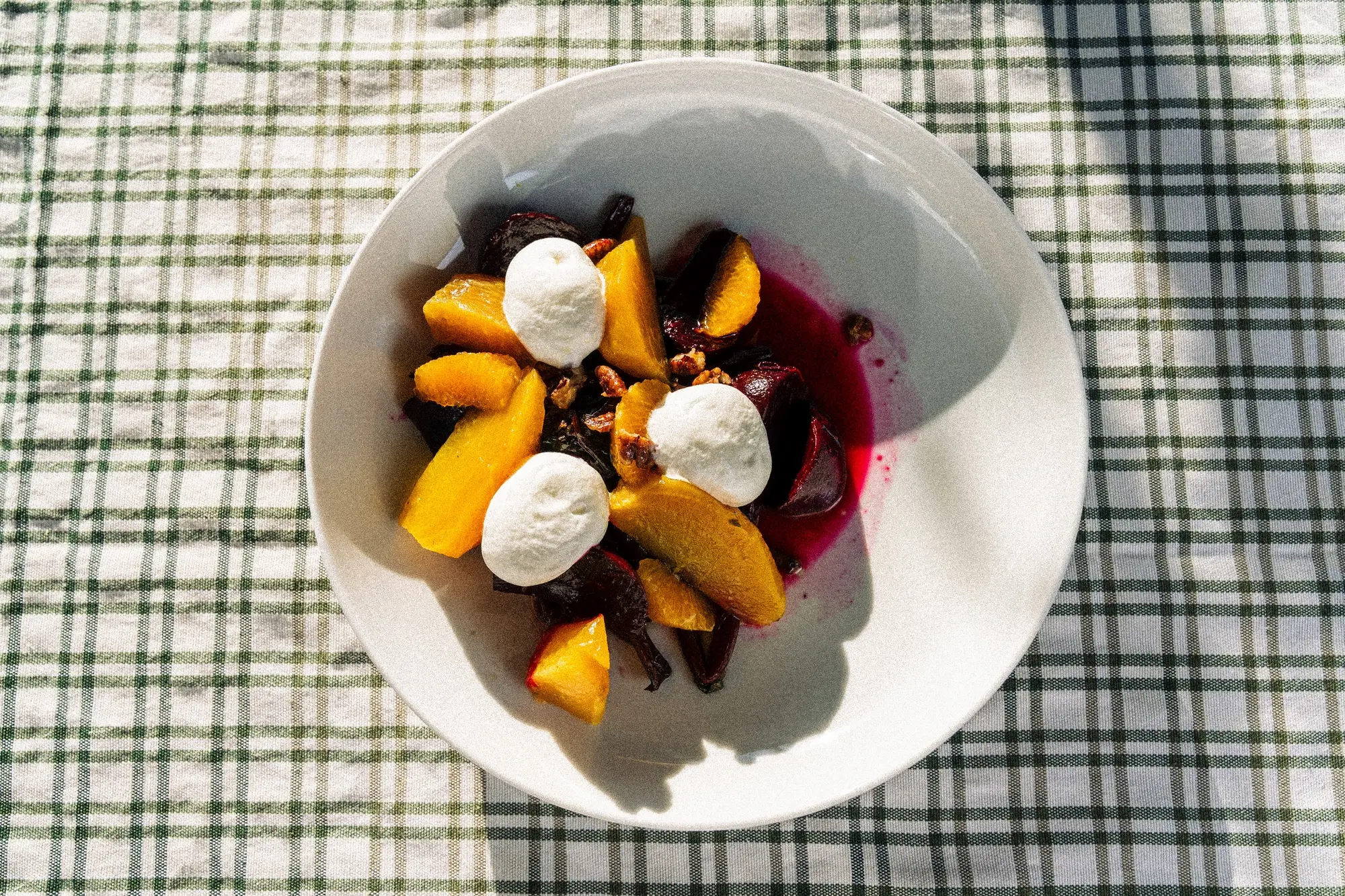 White bowl with beetroot, orange slices, nuts, and dollops of white cream on checked cloth.