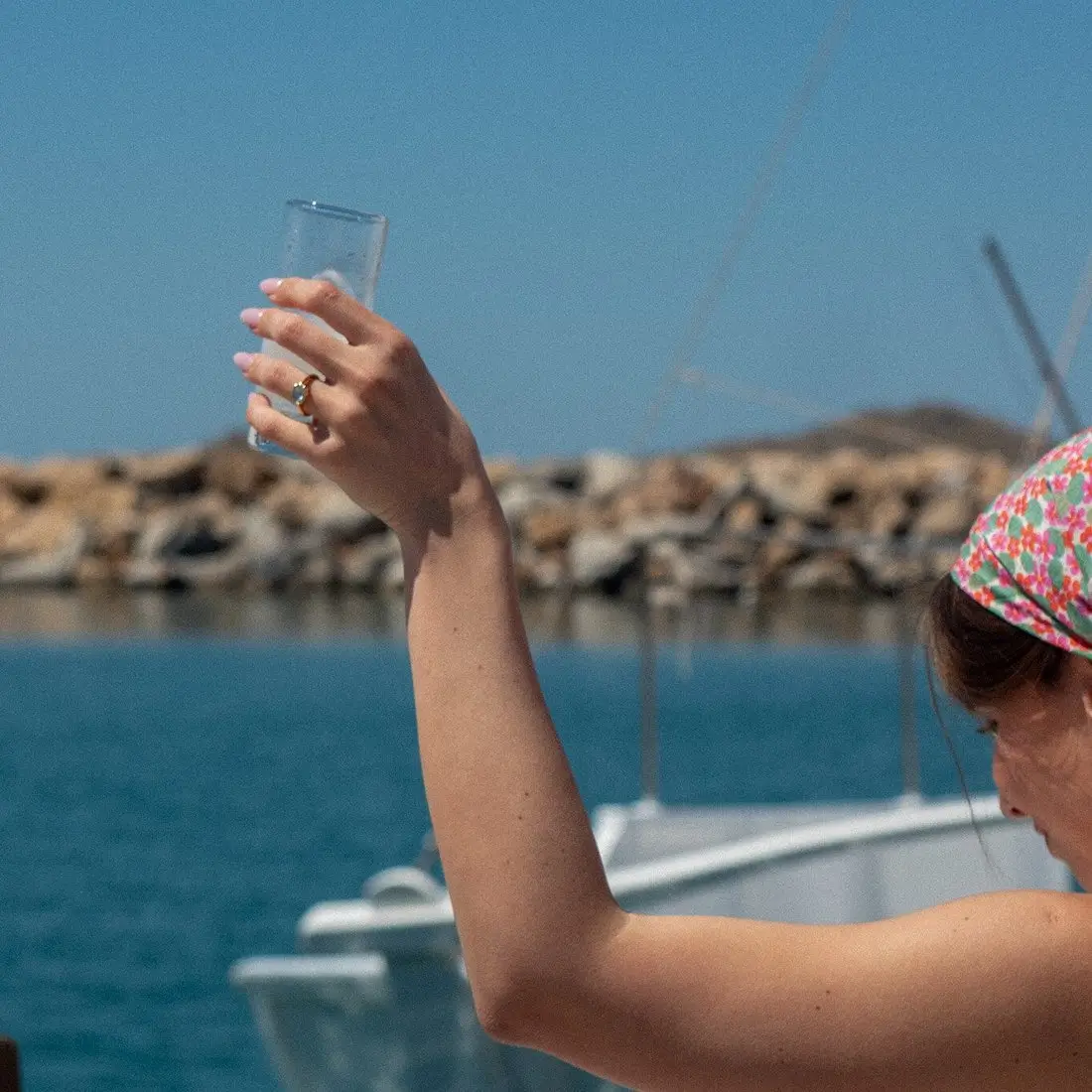 Woman raising a glass while on a boat with rocky shore and calm sea in the background.
